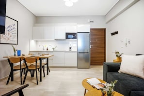 Cuina moderna amb armaris blancs, un frigorífic d'acer inoxidable i un microones. Hi ha una zona de menjador amb cadires de fusta i una taula, i una sala d'estar amb un sofà i una taula de centre.