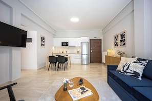 A modern living room with a blue sofa, a coffee table with magazines, a TV, and a kitchen in the background. - FiftySeven Athens - Towards Lycabettus | 203 (Athina)
