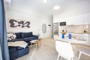 A modern living room with a blue sofa, a dining table with chairs, and a kitchen area with white cabinets and appliances. - FiftySeven Athens - Lycabettus Glimpse | 202 (Athina)
