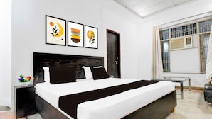A modern bedroom with a large bed, a bedside table, a small table, and a window with an air conditioning unit. - Townhouse Vrindavan Yojna Lucknow (Lucknow)