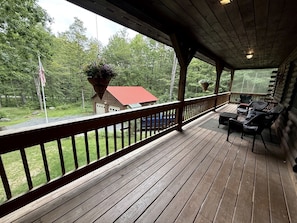 A wooden deck with a railing, outdoor furniture, and a view of a red-roofed building in a forested area. - Peaceful Mountain Escape with Deck and Game Room in Jamaica, Vermont (Jamaica)