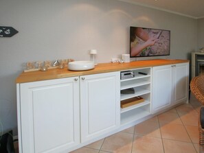 Een keuken met witte kastjes, een houten aanrecht, een spoelbak, een televisie en een rieten stoel.