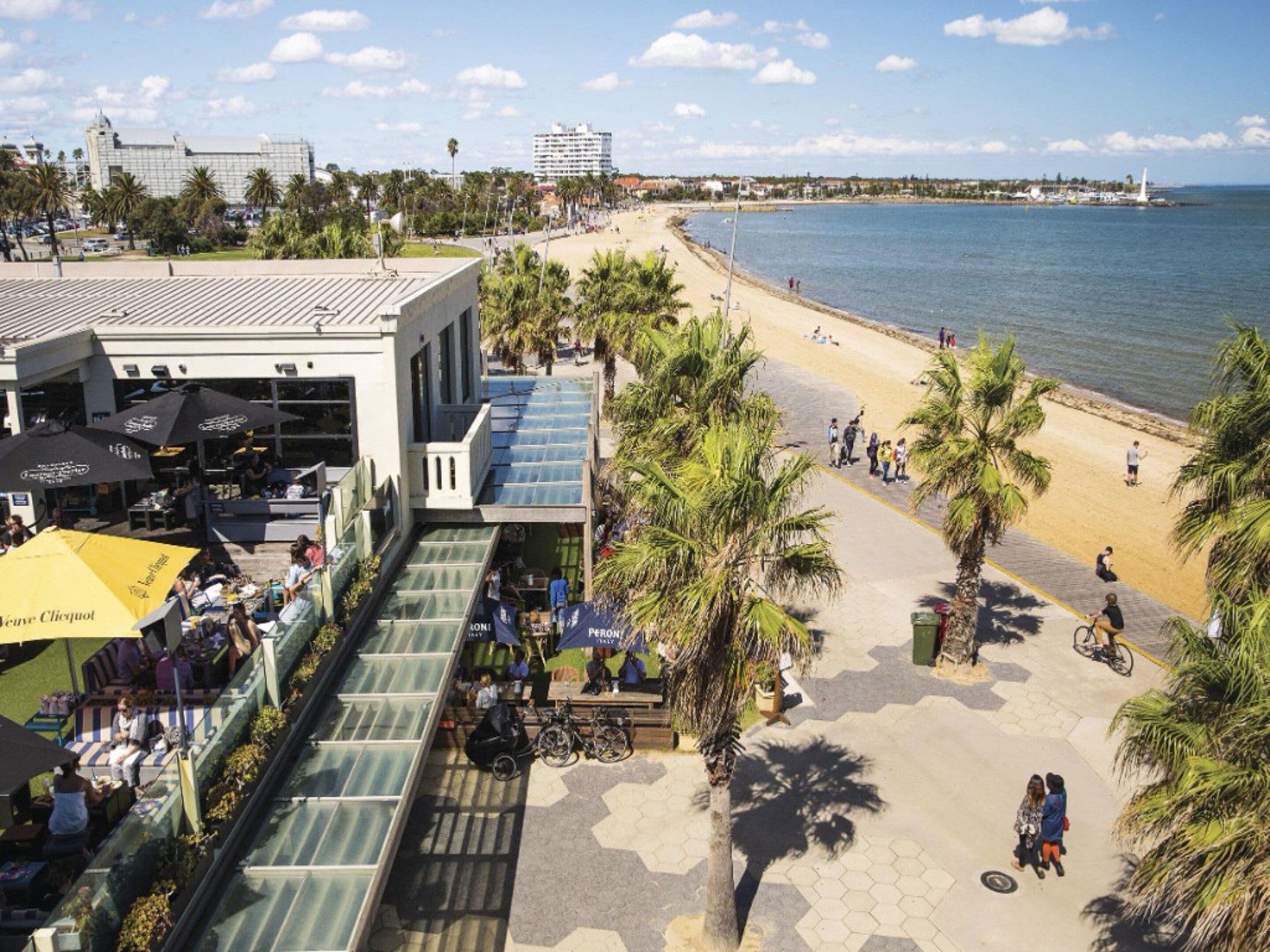 Una passeggiata costiera con spiaggia, palme e una zona con caffè e posti a sedere all'aperto.