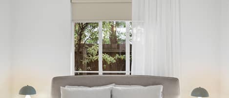 A bedroom with a large bed, two bedside tables with lamps, a window with curtains, and a view of greenery outside.