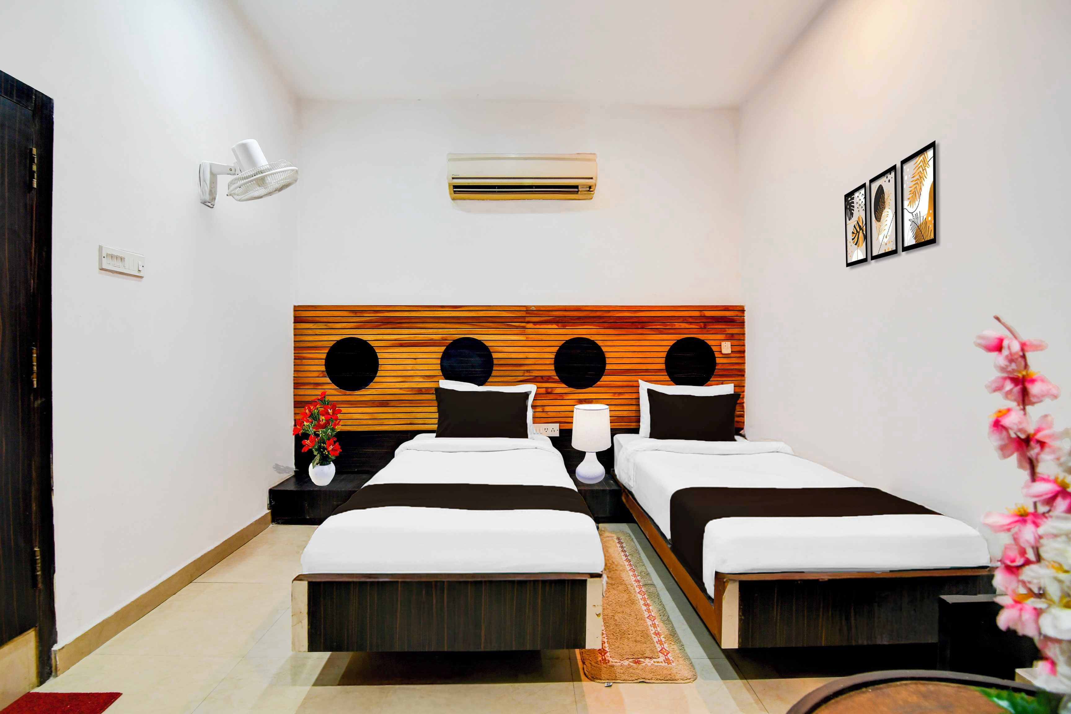 Habitación de hotel con dos camas, cabecera de madera, aire acondicionado instalado en la pared, mesita de noche con lámpara y un pequeño florero con flores.