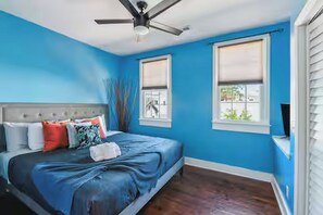 Ein Schlafzimmer mit einem Bett, einem Deckenventilator, zwei Fenstern mit Jalousien und Blick ins Freie.