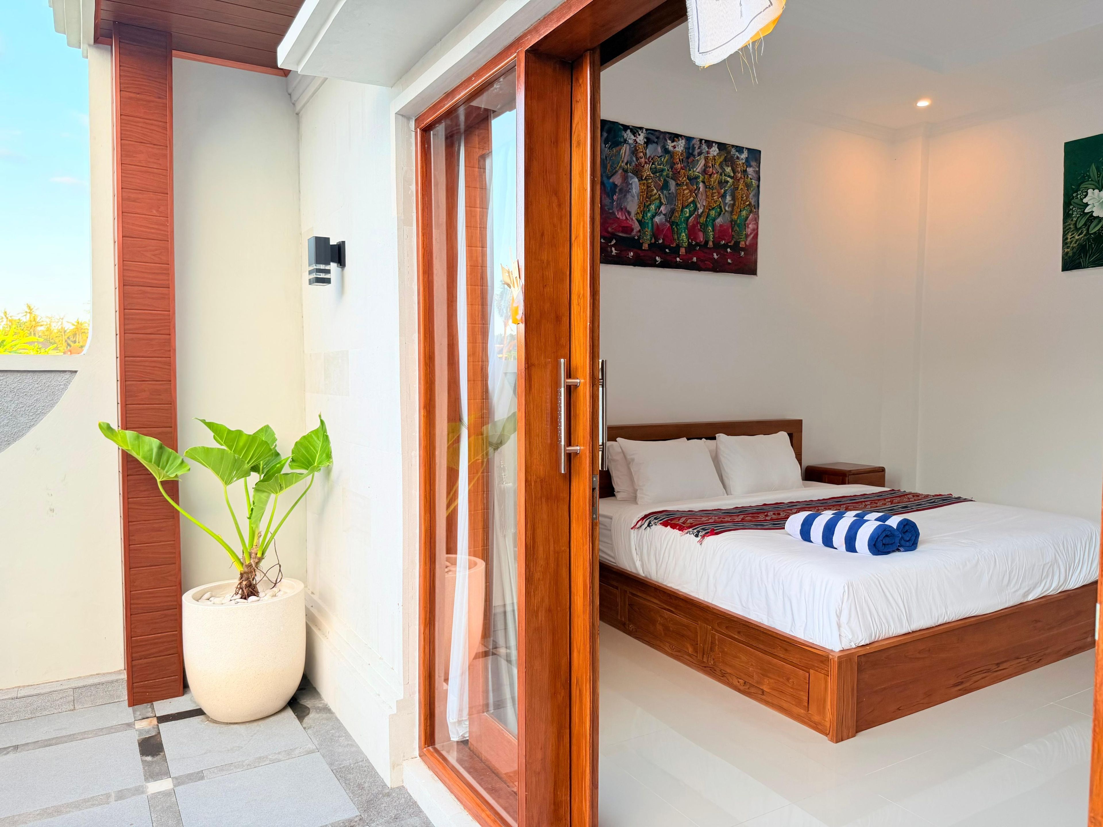 Un dormitorio con cama de madera, ropa de cama blanca, una planta grande en maceta y un amplio ventanal corredero que da a un espacio exterior.