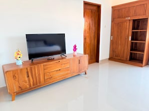 A wooden TV stand with a flat-screen TV, a small vase with flowers, and a pink decorative item. - Villa Romansa Lovina (Buleleng)