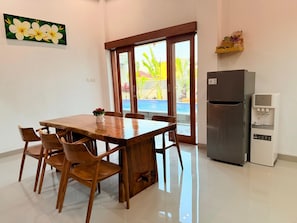 A dining area with a wooden table and chairs, a refrigerator, and a view of a swimming pool through glass doors. - Villa Romansa Lovina (Buleleng)