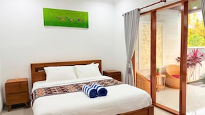 Une chambre d’hôtel avec un lit en bois, du linge de lit blanc, un tableau vert accroché au mur et une porte-fenêtre coulissante donnant sur un espace extérieur agrémenté de plantes.