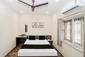 Habitación de hotel con cama, mesitas de noche, ventilador de techo, equipo de aire acondicionado, ventana con barrotes y tres cuadros en la pared.