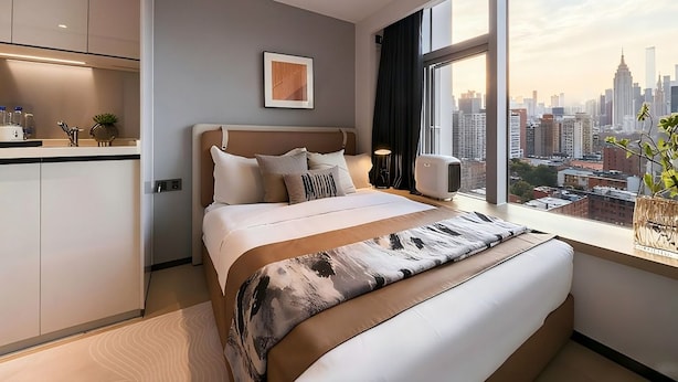Una habitación de hotel moderna con una cama grande, vistas a la ciudad, una kitchenette y un ventanal amplio.