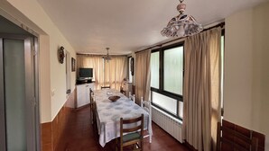 Ein Zimmer mit Esstisch, Stühlen, einem Fernseher, einer Zierleuchte an der Decke und großen Fenstern mit Vorhängen.