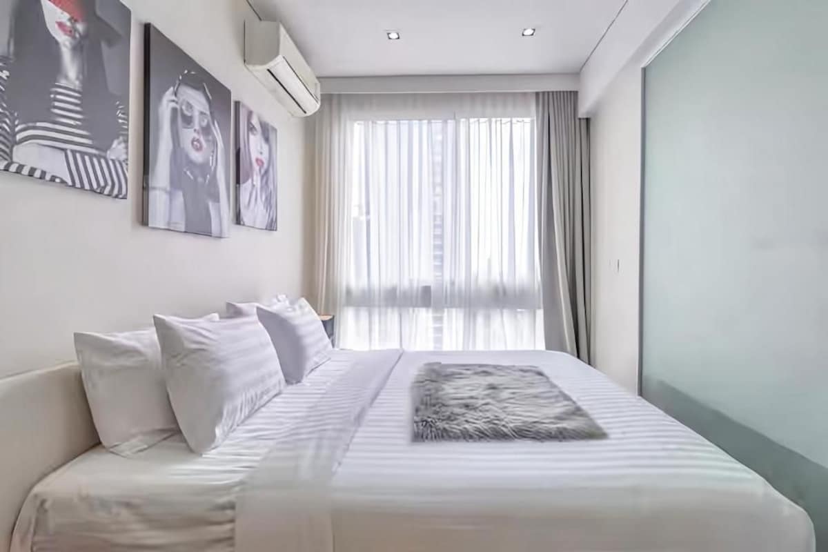 Une chambre à coucher moderne avec un grand lit, du linge de lit blanc et un plaid en fausse fourrure. Trois tableaux encadrés ornent le mur, et une fenêtre est agrémentée de rideaux légers.