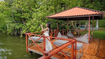 Une terrasse en bois avec une rambarde en métal, un espace salon couvert avec des coussins et une vue sur une étendue d’eau entourée de verdure.
