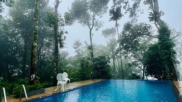 Una piscina azul cristalina rodeada de árboles, con dos sillas blancas y un flotador rojo.