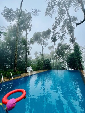 Une piscine d’un bleu éclatant, entourée d’arbres, avec deux chaises blanches et un anneau de bain rouge gonflable.
