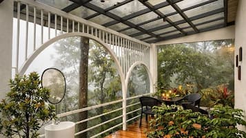 Un espacio al aire libre techado con piso de madera, plantas en macetas, un lavamanos blanco sobre pedestal y una mesa con sillas.