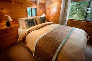A wooden cabin bedroom with a bed, pillows, a lamp, and a window with a view of greenery.