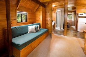 A wooden cabin interior with a sofa, a window, a shower, and a kitchen area.