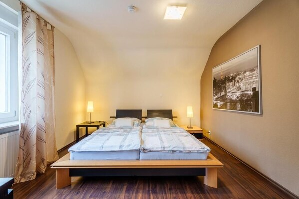 Un dormitorio con una cama, dos mesitas de noche, un mueble auxiliar, una ventana con cortinas y un cuadro colgado en la pared.