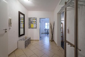Un pasillo con pared blanca, un espejo, un tablón de anuncios, un banco y una puerta que da a otra habitación.