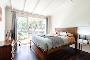 Quarto com uma janela ampla, uma cama de madeira, mesa de cabeceira com abajur e uma cadeira.