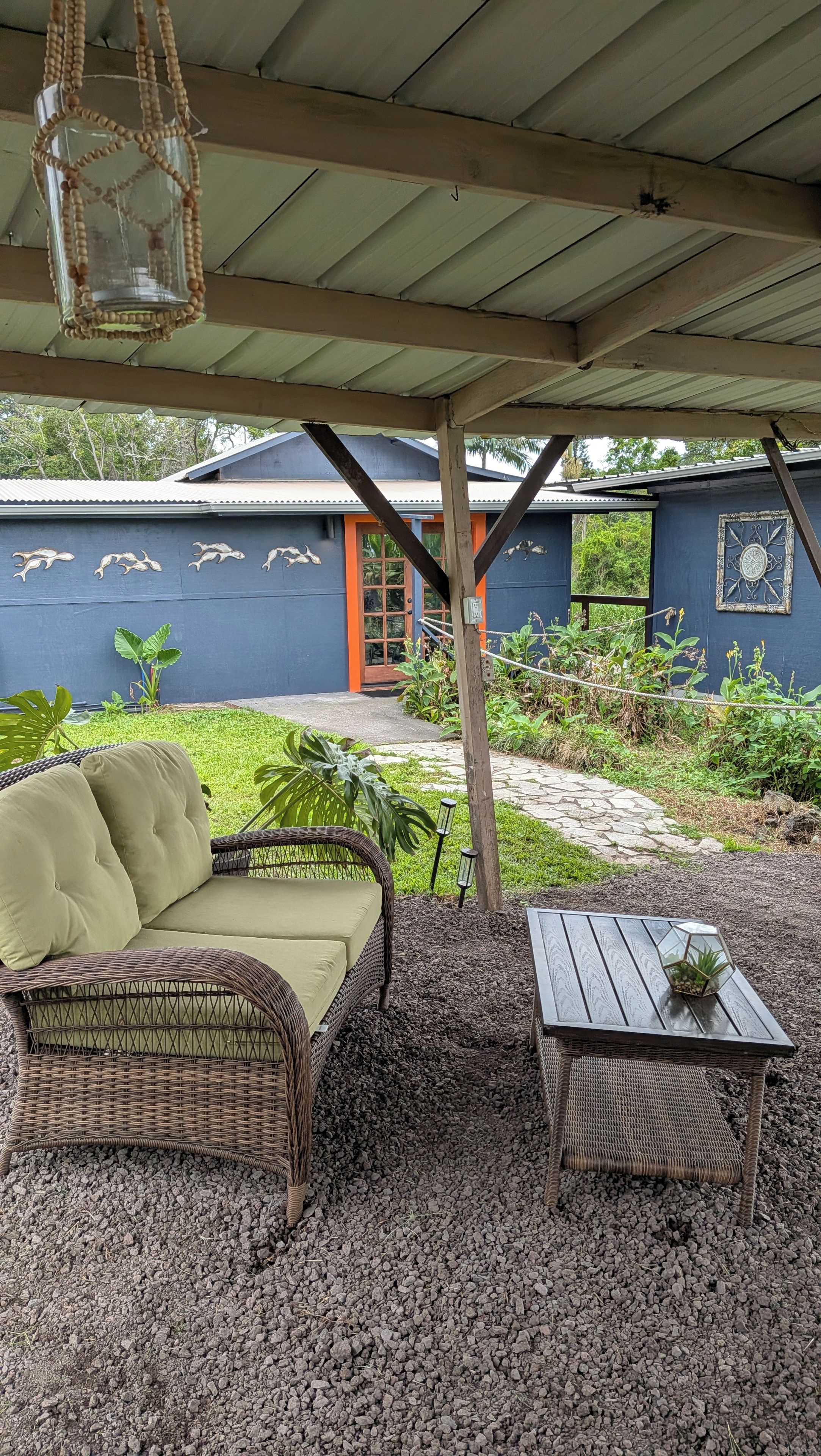 An outdoor seating area with a wicker sofa and a table, surrounded by gravel and plants.