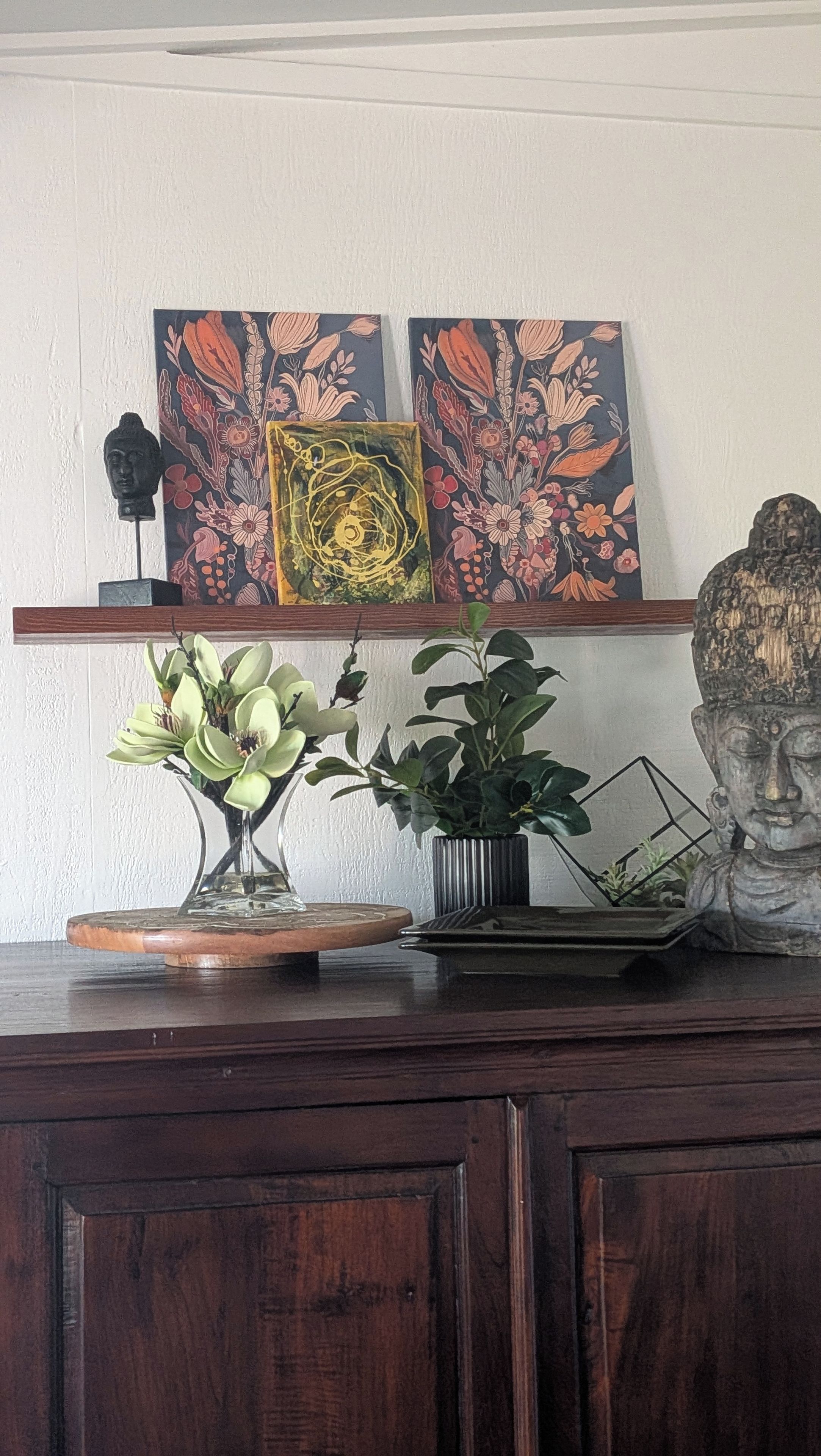 Um armário de madeira com objetos decorativos, incluindo uma escultura de cabeça de Buda, um vaso com flores e duas obras de arte emolduradas.
