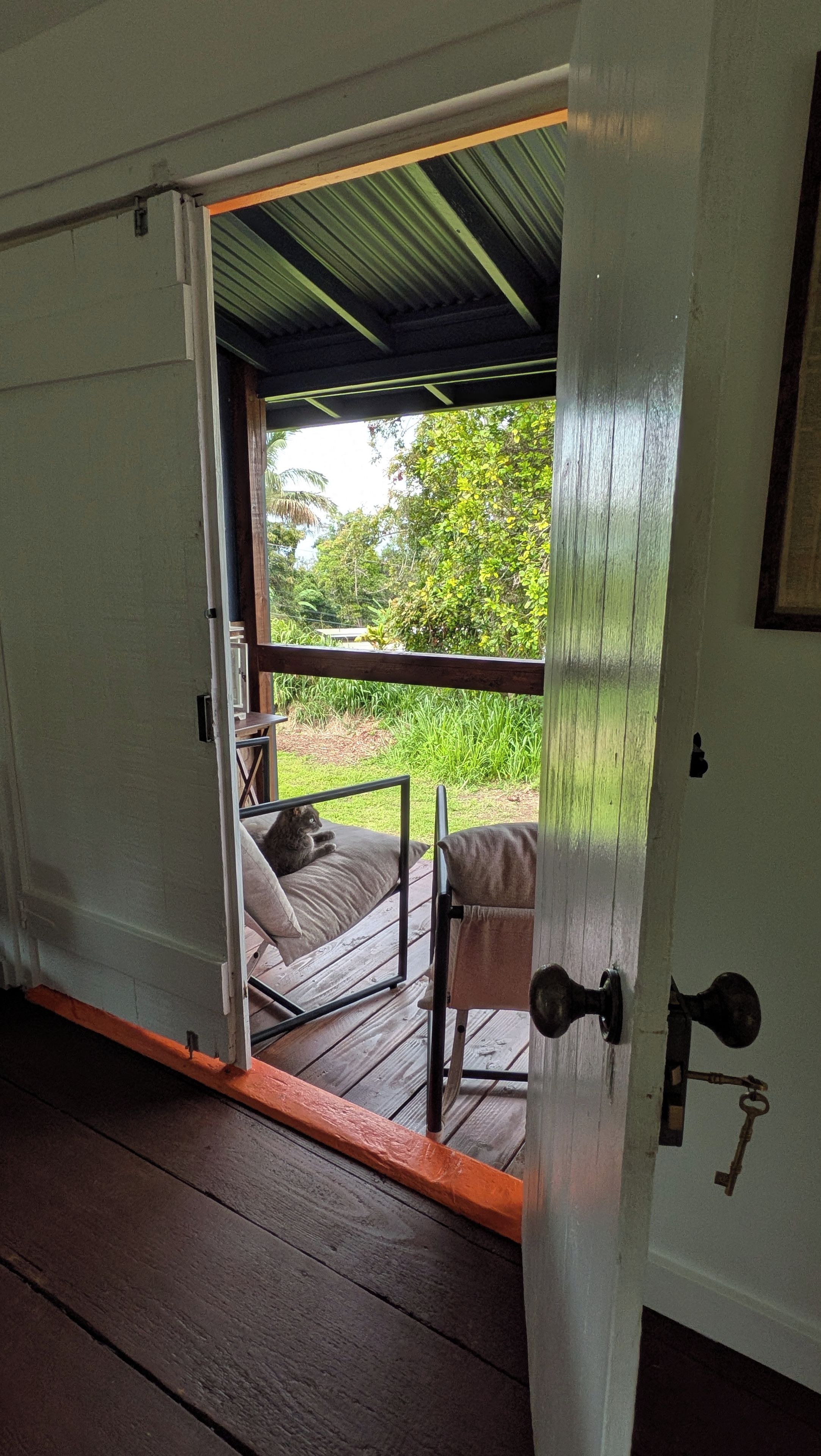 Een geopende deur die leidt naar een veranda met een stoel en een tafel, een houten vloer en uitzicht op groen.