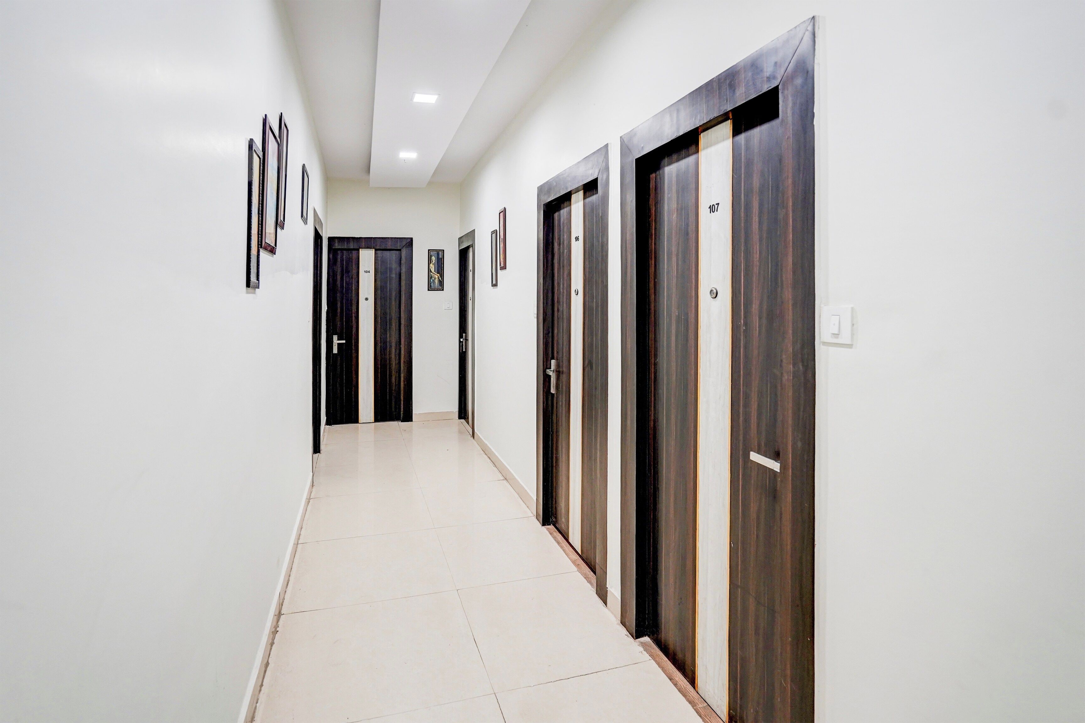 Un pasillo con puertas numeradas del 100 al 107, paredes blancas y cuadros enmarcados a la izquierda.