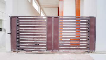 Un cancello metallico con listoni orizzontali e una striscia verticale arancione, che dà accesso a un edificio con facciata bianca e una porta ben visibile.