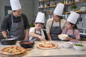 Un groupe de personnes se trouve dans une cuisine, préparant des aliments. On peut voir de la pâte à pizza et divers ingrédients sur le plan de travail.