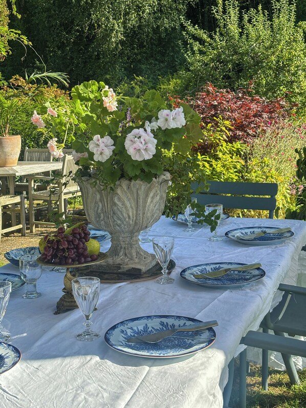 An outdoor dining area with a table set for two, featuring blue and white patterned plates, clear stemware, and a centerpiece of flowers in a stone vase.