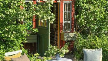 Une construction rustique en bois rouge, dotée d’une porte verte, entourée d’une végétation luxuriante et agrémentée de deux tables blanches avec des chaises.