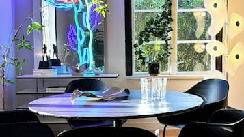 A modern dining area with a round table, black chairs, a neon light sculpture, and a view of greenery through the window.
