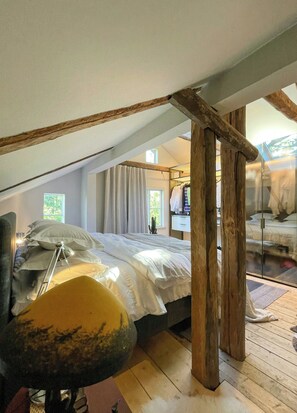 A bedroom with a sloped ceiling, wooden beams, a bed with white bedding, a bedside table, and a window with curtains.