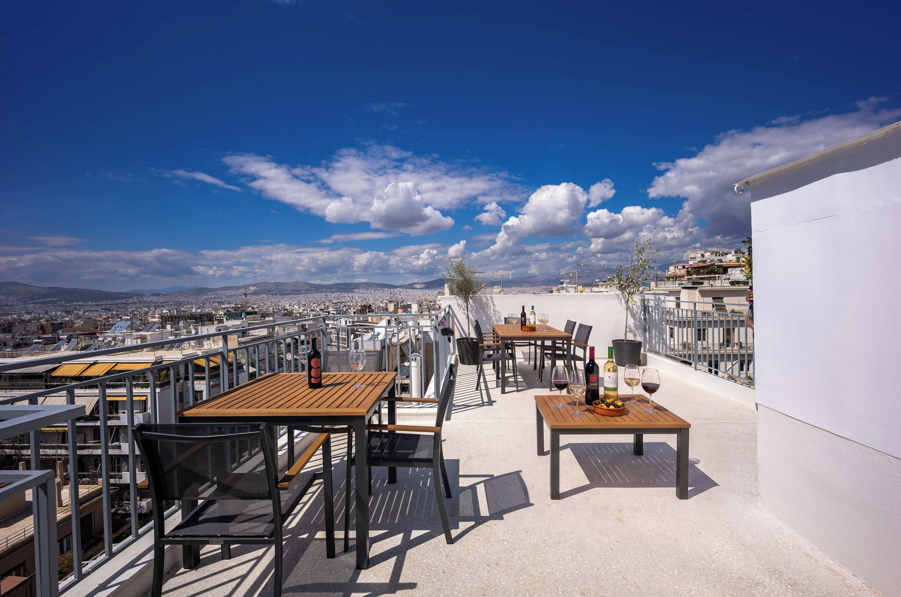 A rooftop terrace with tables and chairs, a view of the city, and a clear sky.