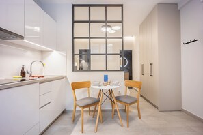 A modern kitchen with white cabinetry, a dining table with two chairs, and a large window with a view of the interior. - FiftySeven Athens - Petite & Practical Studio | B1 (Athina)