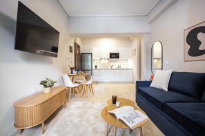 A modern living room with a flat-screen TV, a dark blue sofa, a wooden coffee table with a book, and a dining area with a white table and chairs. - FiftySeven Athens - City Pulse, Balcony | 004 (Athina)