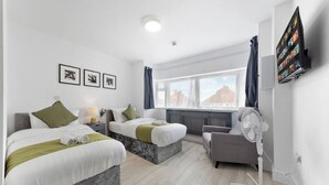 A modern hotel room with two beds, a sofa, a TV, and a window with a view. - Welcome to our modern and spacious 6-bedroom, all en-suite home in **Northolt, West London**, perfect for **up to 12 guests**. Ideal for business travellers, contractors, families, relocation stays, and group trips. (Greater London)