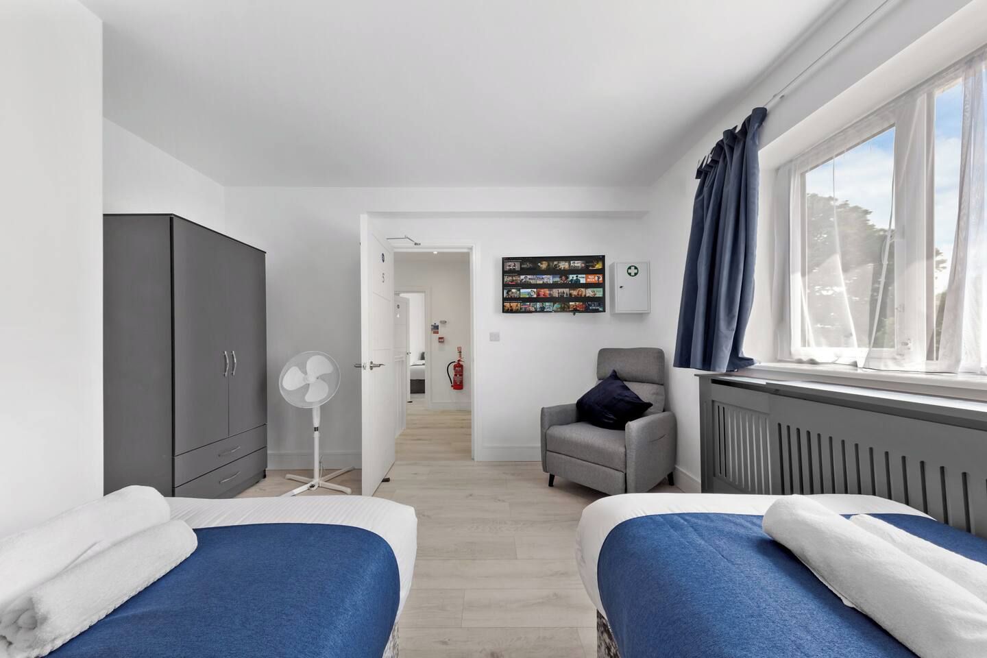A modern hotel room with two beds, a grey wardrobe, a standing fan, a small armchair, a window with curtains, and a wall-mounted photo collage.