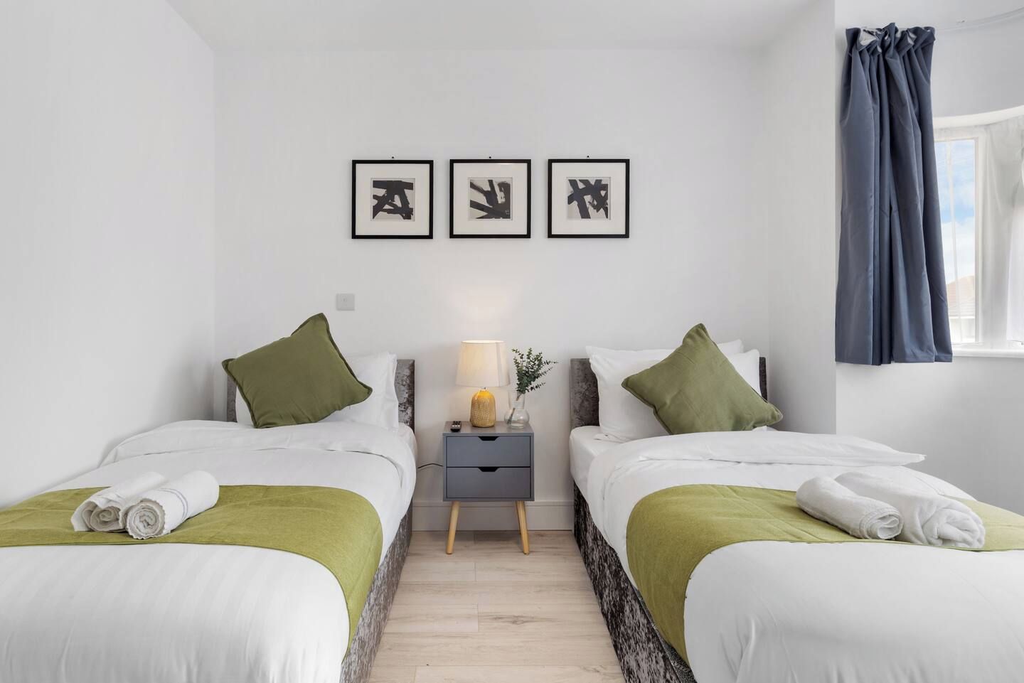Habitación de hotel con dos camas, una mesita de noche con lámpara y tres obras de arte abstracto enmarcadas en la pared.
