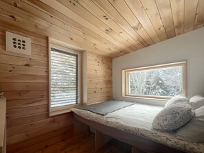 A small, cozy room with wooden walls and a sloped ceiling. There are two windows, one with blinds and one without, providing views of trees. A bed with a patterned bedspread and pillows is against the wall. - The Paradise Cabin w/ Sauna - 10 min to the Falls (Elka Park)