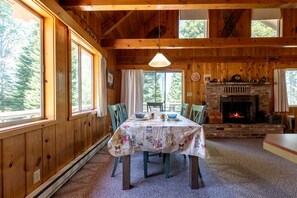 A wooden interior with a dining table set for four, a fireplace, and large windows. - Secluded Island Park Cabin with Hot Tub Near Yellowstone & Henrys Lake, Idaho (Island Park)