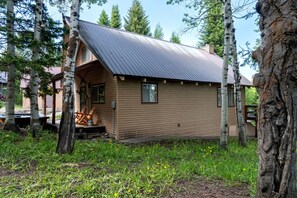 En liten stuga av trä med ett sluttande tak, omgiven av träd och en gräsyta.