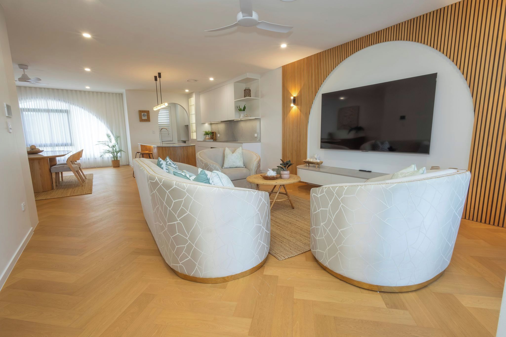 Uma sala moderna com televisão de tela plana, dois sofás brancos com padrões geométricos, um espaço de jantar com mesa e cadeiras de madeira, e uma cozinha ao fundo.