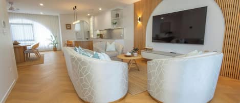Uma sala moderna com televisão de tela plana, dois sofás brancos com padrões geométricos, um espaço de jantar com mesa e cadeiras de madeira, e uma cozinha ao fundo.