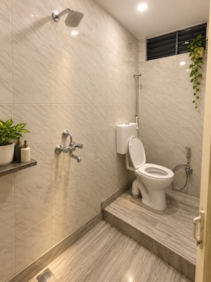 A modern bathroom with a shower, toilet, and a small shelf with toiletries. - Geeta Bhawan Cozy Homestay (Varanasi)