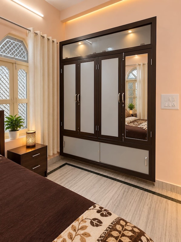 A bedroom with a large wardrobe, a bedside table with a lamp, and a window with curtains. - Geeta Bhawan Cozy Homestay (Varanasi)